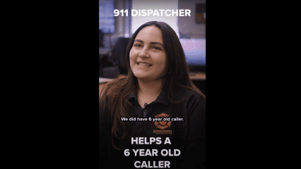 A smiling woman in a black uniform with a fire department emblem sits in a dispatch center, speaking on camera. Behind her are computer monitors and office equipment. Text on the image reads: “911 DISPATCHER” at the top, “We did have 6 year old caller.” in the middle as a subtitle, and “HELPS A 6 YEAR OLD CALLER” at the bottom, highlighting a dispatcher’s role in assisting a young emergency caller.